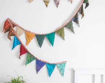 May include: A colorful fabric bunting with triangular flags in various shades of blue, green, red, orange, and yellow. The bunting is hanging on a white wall with a white bed in the background.