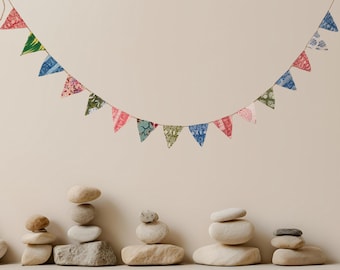 May include: A colorful garland of triangular fabric flags hangs against a neutral background. The flags feature various patterns and colors, including red, blue, green, and pink. Below, several stacks of smooth stones are arranged.