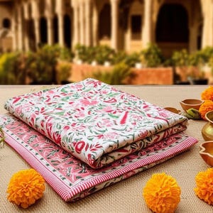 May include: Two folded floral patterned fabric cloths. The top cloth has a white background with red and green floral designs. The bottom cloth is pink with a red border. Orange flowers and small brass pots are also visible.