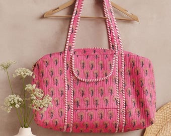 May include: A pink quilted travel bag with a floral pattern, hanging on a wooden hanger. The bag has a rectangular shape with two handles and a small pocket. The trim is white with a thin pink stripe.