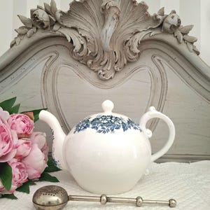 Pode incluir: Um bule de cerâmica branca com um padrão floral azul na parte superior, uma pega curva e um bico. Ao lado do bule encontra-se um infusor de chá em metal prateado com uma cabeça esférica perfurada e um cabo longo e fino.