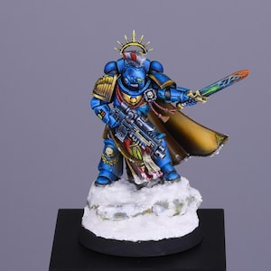 Puede incluir: Una miniatura pintada de azul y dorado de un guerrero espacial marine sosteniendo una espada y un arma. La miniatura está de pie sobre una base nevada.