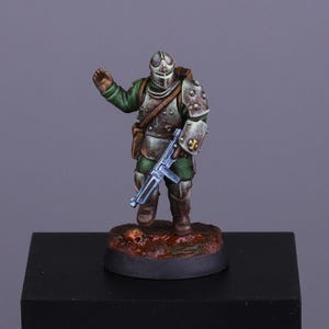 May include: A miniature figurine of a soldier in green and silver armour, holding a blue submachine gun. The figure stands on a brown base with a skull. The background is a solid grey.