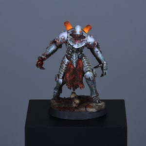 May include: A painted miniature figurine of a metallic armored warrior with a helmet, glowing orange exhaust vents on the shoulders, and a red chainmail skirt. The figure stands on a base featuring small skulls and dark, textured ground details.