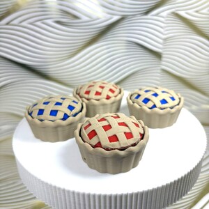 May include: Four miniature pie replicas featuring beige crusts with a lattice top design. Two pies have a bright red filling visible through the lattice, and two pies have a vibrant blue filling. Each pie has a scalloped edge along the base.