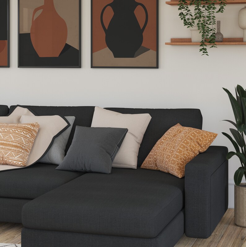 May include: A black sectional sofa with a chaise lounge, featuring a variety of throw pillows in beige, grey, and orange with geometric patterns. The sofa is positioned in front of a white wall with framed artwork and a shelf with a potted plant.