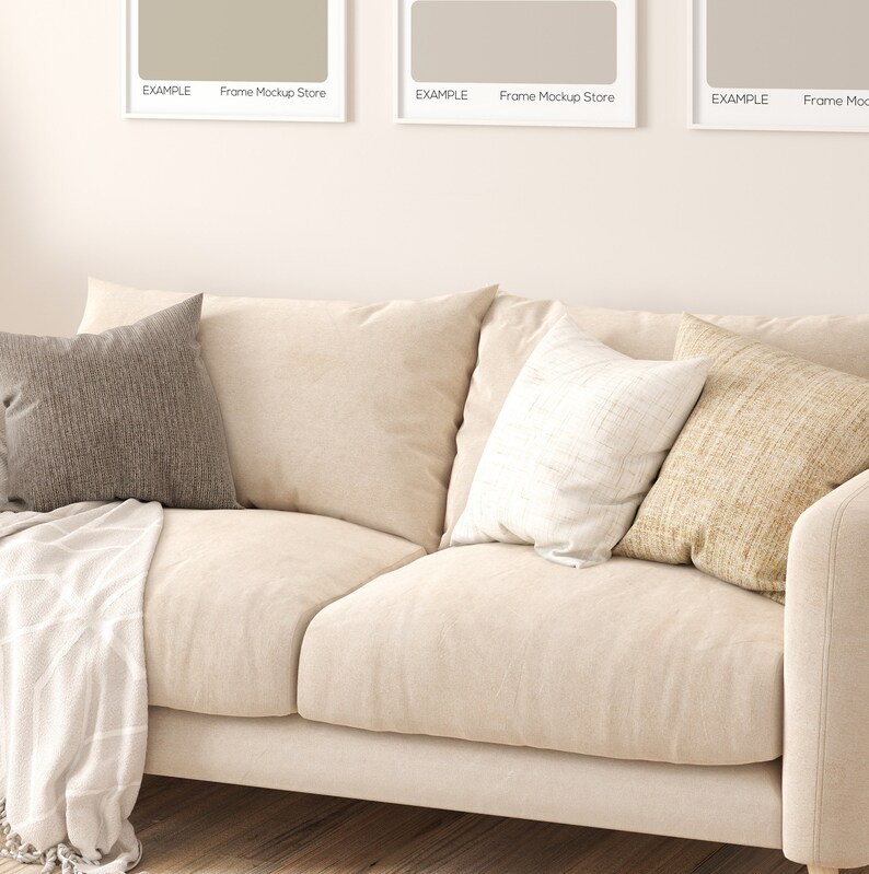 May include: A beige sofa with three pillows. The pillows are brown, white, and beige. The sofa is in front of a white wall with two framed pictures above it.