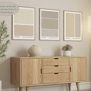 May include: Three framed art prints with neutral color block designs hang above a wooden cabinet. The prints are in light brown frames. A lamp and a potted plant are also in the room. The text "Add your artwork using smart layers in Photoshop" is in the image.