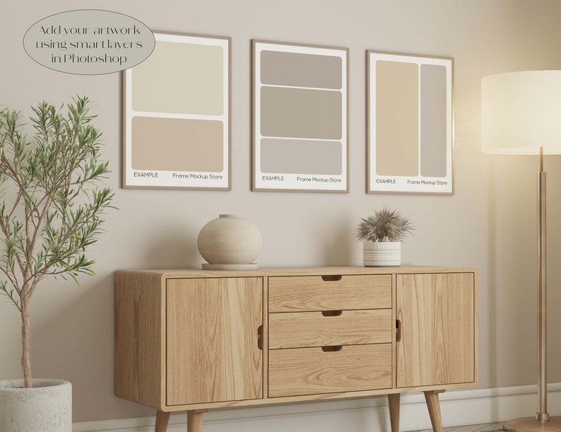 May include: Three framed art prints with neutral color block designs hang above a wooden cabinet. The prints are in light brown frames. A lamp and a potted plant are also in the room. The text "Add your artwork using smart layers in Photoshop" is in the image.