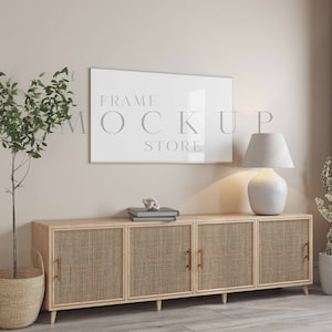 May include: A white framed picture with the text "FRAME MOCKUP STORE" hanging on a beige wall above a light wood cabinet with three woven doors. A white lamp with a white lampshade sits on the cabinet. A small potted plant is on the right side of the cabinet.