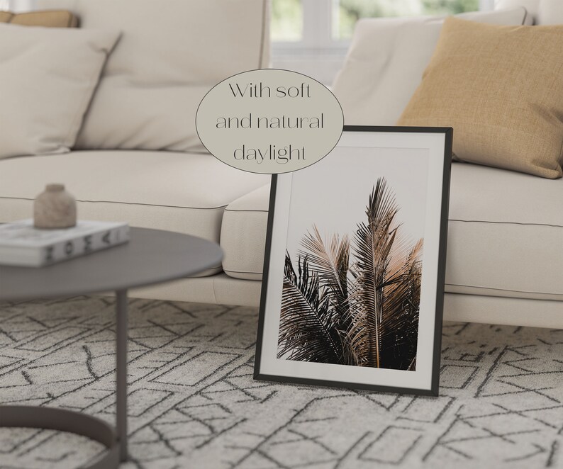 May include: A black framed print of palm trees with a white background. The print is sitting on a white couch with a beige throw pillow. The couch is in a living room with a coffee table and a patterned rug.