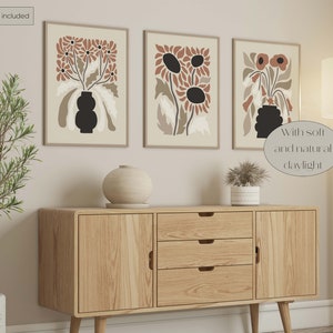 May include: Three framed floral art prints with a beige background and brown and black flower designs. The prints are displayed above a wooden cabinet with a round vase and a potted plant. The text "Artwork not included" is in the top left corner.