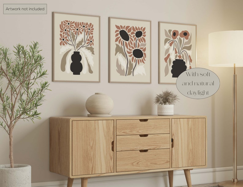 May include: Three framed floral art prints with a beige background and brown and black flower designs. The prints are displayed above a wooden cabinet with a round vase and a potted plant. The text "Artwork not included" is in the top left corner.