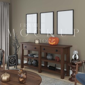 Puede incluir: Tres cuadros negros enmarcados cuelgan de una pared marrón sobre una consola de madera marrón oscuro con dos cajones y dos estantes. La mesa está decorada con una calabaza tallada, una calabaza rayada en blanco y negro, una calabaza negra y naranja, una maceta negra, una figurita de elefante marrón y dos libros. Un letrero en blanco y negro con el texto "ALIEN" está en el suelo a la izquierda de la mesa.