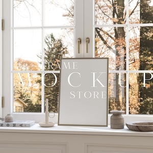 May include: A gold frame mockup with the text "FRAME MOCKUP STORE" on a white windowsill with a book, candle, and decorative items. The window is behind the frame and shows a view of trees with fall foliage.