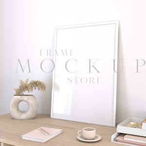 May include: A white picture frame mockup on a wooden desk with a vase of dried flowers, a notebook, a pen, and a cup of coffee. The frame is blank and ready for your design.