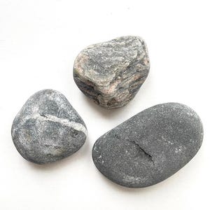 Puede incluir: Tres piedras lisas de color gris, de diferentes tamaños y formas. La piedra más grande tiene un patrón de mármol rosa y gris.