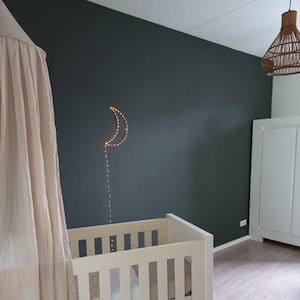 Può includere: Un lettino bianco con un baldacchino bianco appeso sopra. La parete dietro il lettino è dipinta di un verde scuro e ha una luce a forma di mezzaluna con luci a stringa che pendono da essa.