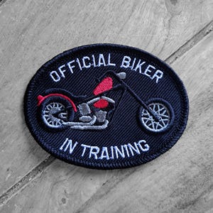 May include: Navy blue oval patch featuring a motorcycle design with the text "OFFICIAL BIKER IN TRAINING". The motorcycle is depicted in black, red, and grey. The patch is set against a wooden background.