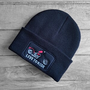May include: A dark navy beanie hat with a rectangular patch. The patch displays a red and silver motorcycle graphic and the words "LIVE TO RIDE" in white. The hat is made of a knitted material.