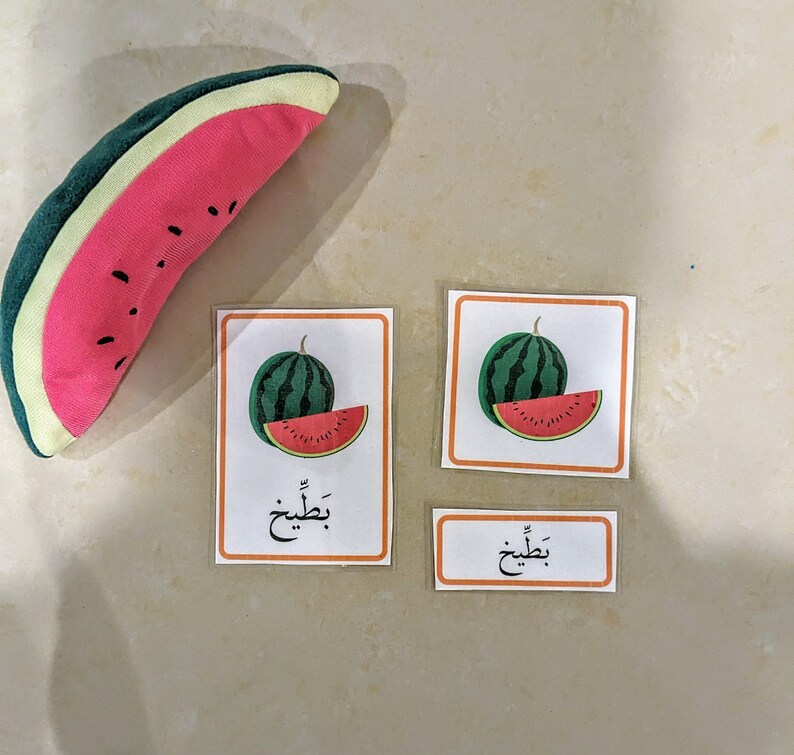 May include: Three flashcards with images of watermelon slices and the Arabic word for watermelon, "بطيخ", written on each card. The cards are on a light beige surface.