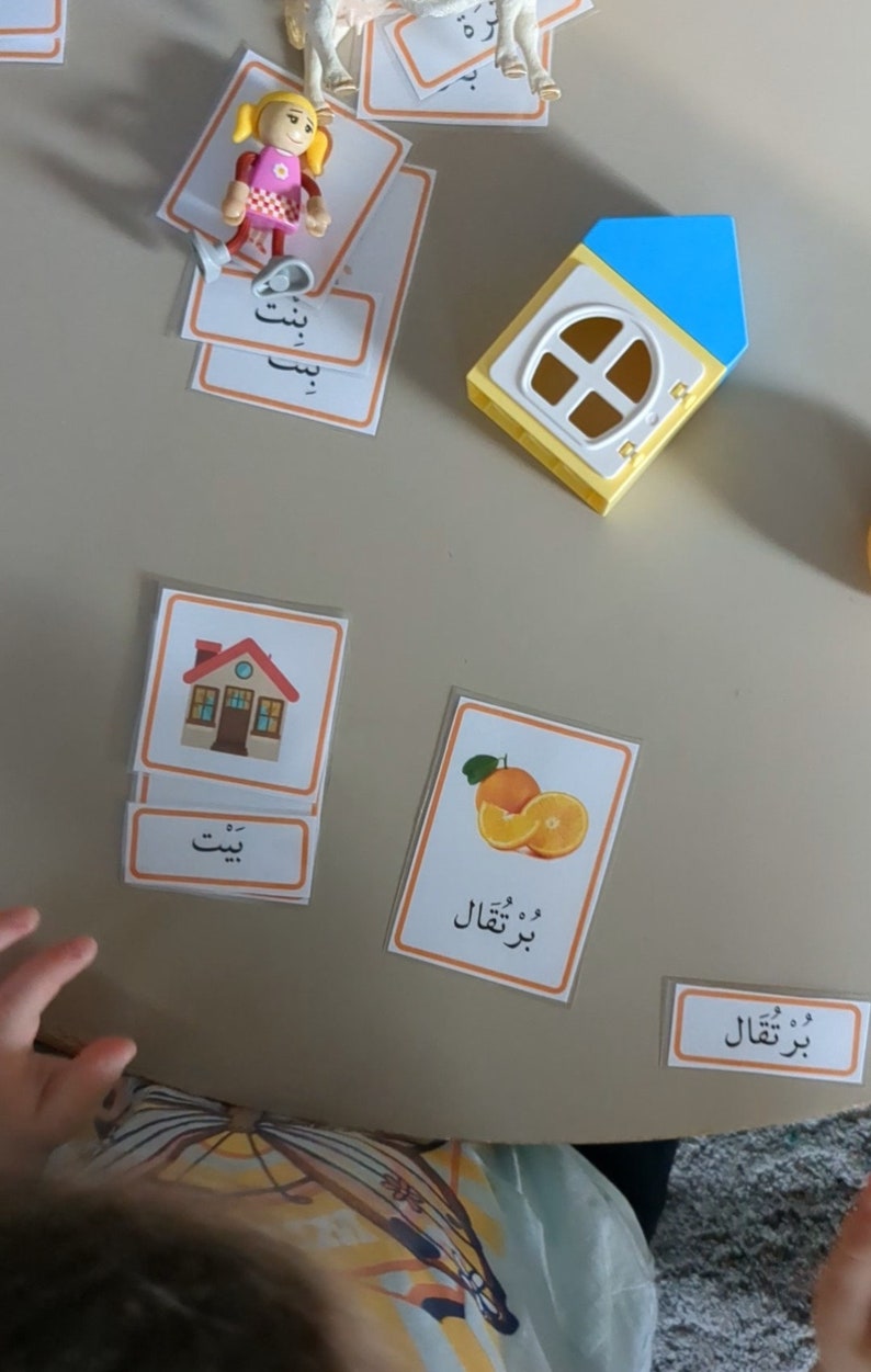 May include: A set of colorful flashcards with Arabic words and pictures. The cards feature a house, an orange, and a girl. The words on the cards are "بيت" (house), "برتقال" (orange), and "بنت" (girl).