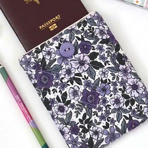Puede incluir: Una funda para pasaporte con un estampado floral en tonos morados, grises y blancos. La funda se cierra con un botón morado. Un pasaporte es parcialmente visible y un lápiz está en primer plano.
