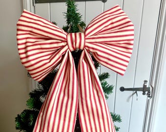 May include: A large decorative bow made of fabric with a red and cream-coloured vertical striped pattern. The bow features two wide loops at the top and two long, hanging tails extending downward, positioned at the top of a green artificial tree.