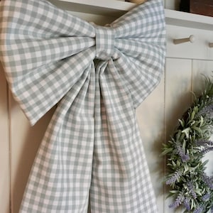 May include: A large decorative bow made of fabric featuring a light sage green and white gingham check pattern. The bow has two wide, long tails hanging downwards and a central knot, creating a structured and symmetrical appearance.