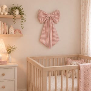 May include: A large decorative fabric bow featuring a pink and white vertical striped pattern. The bow is mounted on a wall and consists of two wide loops at the top with two long, pointed tails hanging downward, creating a symmetrical shape.