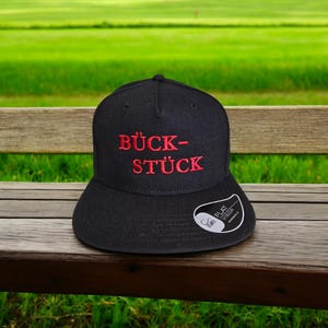 May include: A black baseball cap with a red embroidered phrase "BÜCK-STÜCK" on the front. The cap has a flat brim and a white label on the side that says "FLAT VISOR".