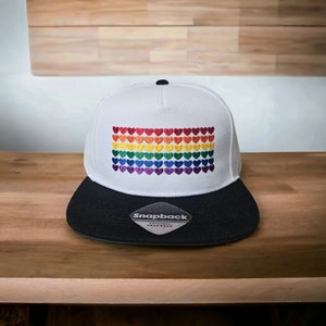 Può includere: Cappellino da baseball bianco e nero con un design a forma di cuore arcobaleno e il testo "Snapback Original Headwear" sul visore.