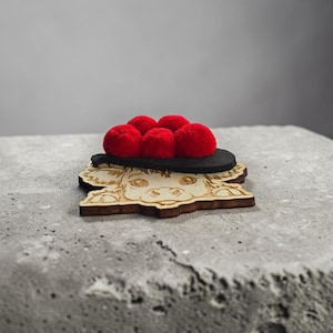 Può includere: Un sottobicchiere in legno a forma di mucca, con un design tagliato al laser. Ha un cappello di feltro nero con cinque pompon rossi. Il sottobicchiere è realizzato in legno chiaro e poggia su una superficie grigia.