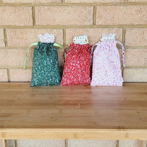 May include: Three festive drawstring gift bags in green, red, and pink, each with a holiday pattern. The green bag says "Magic Something", the red bag says "Cookies for Santa", and the pink bag says "Happy Holidays".
