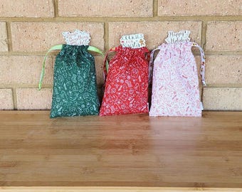 May include: Three festive drawstring gift bags in green, red, and pink, each adorned with a holiday-themed pattern. The green bag says "Magic Something", the red bag says "Cookies for Santa", and the pink bag says "Happy Holidays".