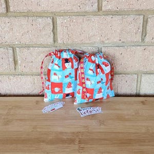 May include: Two festive drawstring gift bags with a Christmas pattern featuring snowmen, gnomes, and mugs on a blue backdrop. The bags have red and white ribbon drawstrings. Small gift tags with text are in front.