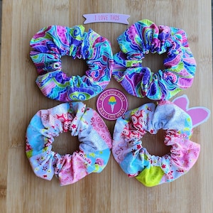 May include: Four colourful fabric hair scrunchies on a wooden surface. Two scrunchies have a paisley pattern in shades of pink, purple, and green. The other two scrunchies have a patchwork pattern in shades of pink, blue, and green. One of the scrunchies has a pink bunny ear tag attached. The text "I love this" is on a pink banner above the scrunchies. The text "Beat the Heat" is on a pink circle with a pink ice cream cone graphic.