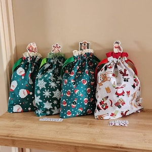May include: Four green Christmas gift bags with different festive patterns. The bags have red ribbon ties and are decorated with festive stickers. The bags are sitting on a wooden shelf.