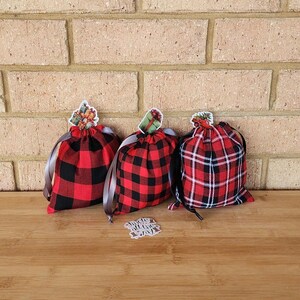 May include: Three red and black buffalo check drawstring bags with decorative gift tags. The bags are tied with brown ribbon and sit on a wooden surface against a brick wall. One tag reads "Jingle all the way."