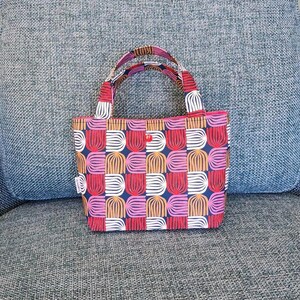 May include: A small tote bag with a colorful geometric pattern of white, pink, orange, and blue shapes. The bag has matching handles and a red button closure. The bag is sitting on a blue couch.
