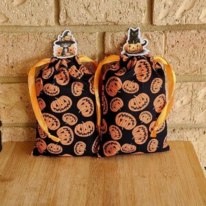 May include: Two black Halloween treat bags with orange pumpkin designs and orange ribbon handles. Each bag has a decorative sticker of a black cat wearing a witch hat. The bags are on a wooden surface.