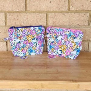 May include: Two zippered cosmetic bags with a floral and Snoopy print. The bags feature a colorful pattern of flowers in shades of pink, purple, yellow, and blue. The bags are sitting on a wooden surface.