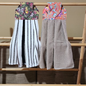 May include: Two kitchen towels with floral fabric accents hanging on a wooden rack. The towel on the left has black and white stripes, while the towel on the right is a solid beige color.