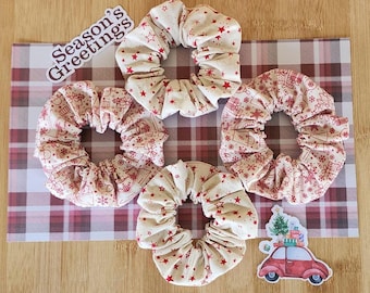 May include: Four Christmas-themed scrunchies in red and white fabric with a plaid background. The scrunchies feature a variety of holiday designs, including stars, houses, and a car with a Christmas tree on top. The text "Season's Greetings" is visible in the top left corner of the image.