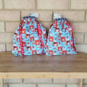 May include: Two drawstring gift bags with a festive winter pattern. The bags feature a repeating design of snowmen, trees, and mugs on a light blue background with red squares. Red ribbon with white snowflakes accents the bags. The words "Just Believe" and "Jingle all the way" are on the tags.