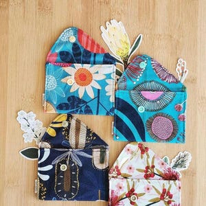 May include: Four fabric pouches with floral patterns in blue, white, and red, arranged on a wooden surface. Each pouch has a unique design, with attached decorative floral accents. The pouches are small and appear to be handmade.