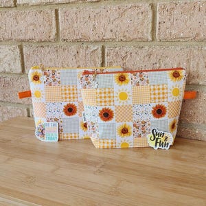 May include: Two cosmetic bags with a patchwork design featuring sunflowers, yellow gingham, and floral patterns. The bags have orange zip pulls and the text "Sun & Fun" on one bag. The other bag has the text "Be the reason someone smiles today".