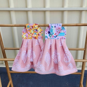 May include: Two pink hand towels with white seashell patterns hang on a wooden rack. The top of each towel has a colourful patterned fabric with text and images of stars, shells, and other sea-themed elements.