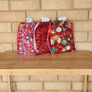 May include: Three festive drawstring bags with holiday-themed patterns. One bag features owls and trees, another has text that says "Peace, Joy, Hope, Light", and the third has Christmas ornaments. Each bag has a decorative ribbon.