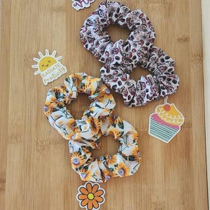 Peut inclure: Trois chouchous avec des motifs différents. Un chouchou a un motif de tournesol, un autre a un motif de cupcake et le dernier a un motif de différentes formes colorées. Les chouchous sont sur une surface en bois avec des autocollants qui disent "Good Vibes", "Radiate Positivity" et une fleur jaune.
